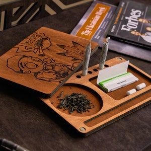 May include: A rectangular wooden rolling tray with a dark brown finish featuring laser-engraved cartoon characters on the lid. The tray includes a circular compartment for loose herbs, slots for two pre-rolled items, and a space for rolling papers.