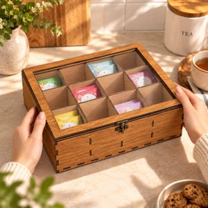 May include: A rectangular wooden tea box with a clear glass lid and a metal latch. The box features twelve individual compartments, some of which contain colourful tea bags. The wood has a natural brown grain and visible joinery details.