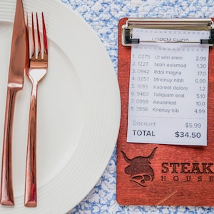 May include: A white plate with a rose gold knife and fork set next to a wooden clipboard. The clipboard has a receipt with itemized costs and the text "STEAK HOUSE". The total is $34.50.