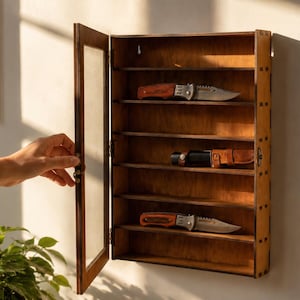 May include: A rectangular wooden wall-mounted display case with a hinged glass door. The interior features six horizontal shelves, with three knives displayed on the shelves. The case has a natural wood finish and visible joinery on the side panels.