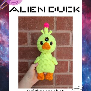 Pode incluir: Um pato alienígena de crochê verde limão feito à mão com três olhos, bico e pés laranja e uma crista rosa. O texto "ALIEN DUCK" está acima do pato e o nome de usuário do Instagram "@rightocrochet" está abaixo.