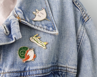May include: A denim jacket with three enamel pins. The pins are of a sea creature, a snail, and a jellyfish. The pins are all gold colored with colorful details.