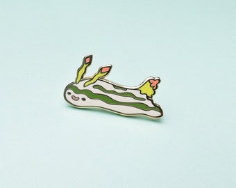 May include: A gold-colored enamel pin featuring a cartoon sea slug with green and white stripes, yellow and orange antennae, and a pink tail.