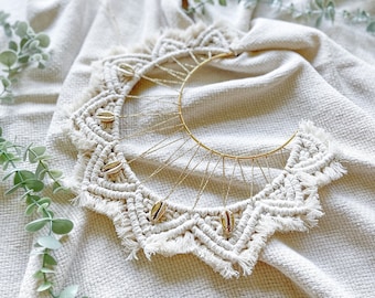 Op de afbeelding: Een witte macramé wandhanger in de vorm van een halve maan met een gouden metalen ring en kleine schelpen als accenten.
