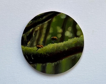 May include: A circular painting depicting a forest scene with two small orange mushrooms growing on a mossy branch. The background is a blurred green and brown forest with dappled sunlight.