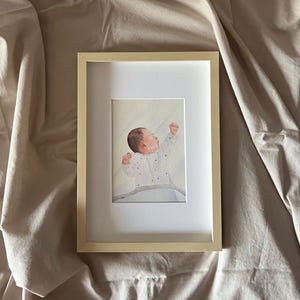 May include: A framed watercolour painting of a baby in a white onesie with star patterns, arms raised. The artwork is set within a light wood frame and a white mount, resting on a beige fabric surface.