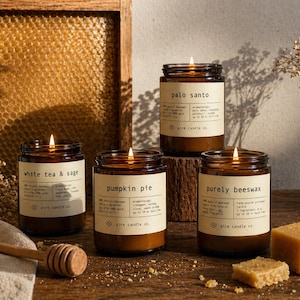 May include: Four lit, amber-colored glass jar candles with white labels, featuring scents like "white tea & sage," "pumpkin pie," "purely beeswax," and "palo santo." A wooden honey dipper and beeswax block are also visible.
