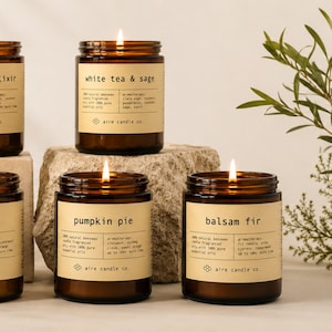 May include: Five amber glass jar candles with lit wicks and beige labels. The labels feature the text aire candle co. and scent names: Lemongrass Elixir, White Tea & Sage, Palo Santo, Pumpkin Pie, and Balsam Fir, listing ingredients and burn times.