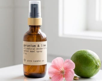 May include: An amber glass spray bottle with a black nozzle and a beige label reading geranium & lime all-natural deodorizing toilet bowl spray 2 oz aire candle co. A pink geranium flower and a whole green lime sit beside the bottle.