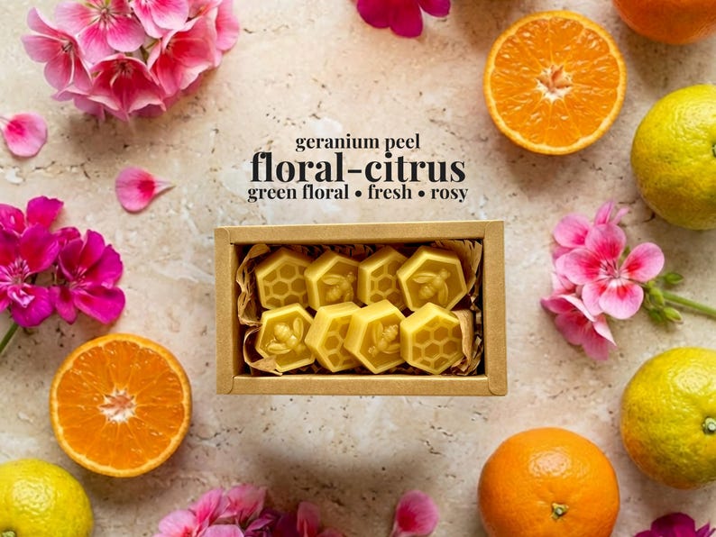 May include: A rectangular cardboard box containing eight yellow hexagonal wax melts, each embossed with bee or honeycomb patterns. The text above the box reads: geranium peel, floral-citrus, green floral, fresh, rosy. Surrounded by pink flowers and citrus fruits.