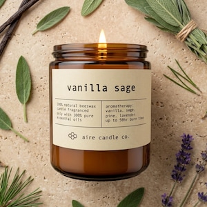 May include: A lit candle in an amber glass jar with a beige label reading vanilla sage, 100% natural beeswax, and aire candle co. The label lists aromatherapy ingredients including vanilla, sage, pine, and lavender with a 50hr burn time.