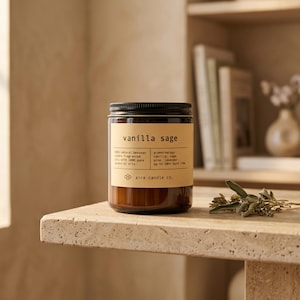 May include: A brown glass candle jar with a black lid and a beige label featuring the text vanilla sage, 100% natural beeswax, aromatherapy, and aire candle co. The candle is positioned next to a small sprig of dried sage leaves on a stone surface.