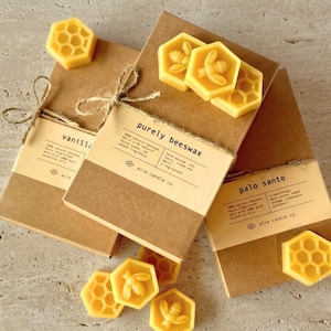 Yellow beeswax wax melts in hexagonal shape sitting atop eco-friendly packaging. Half of the wax melts are shaped like a honeybee and the other half are honeycomb. Featured scents include Purely Beeswax, Palo Santo and Vanilla Sage.