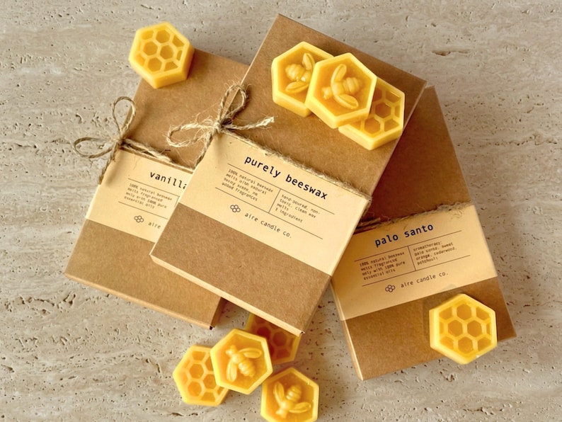 Yellow beeswax wax melts in hexagonal shape sitting atop eco-friendly packaging. Half of the wax melts are shaped like a honeybee and the other half are honeycomb. Featured scents include Purely Beeswax, Palo Santo and Vanilla Sage.
