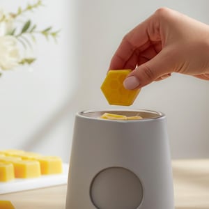 May include: A matte light gray ceramic wax warmer with a circular indentation on the front. A bright yellow hexagonal wax melt with a honeycomb pattern is being placed into the top basin, which contains additional yellow wax pieces.