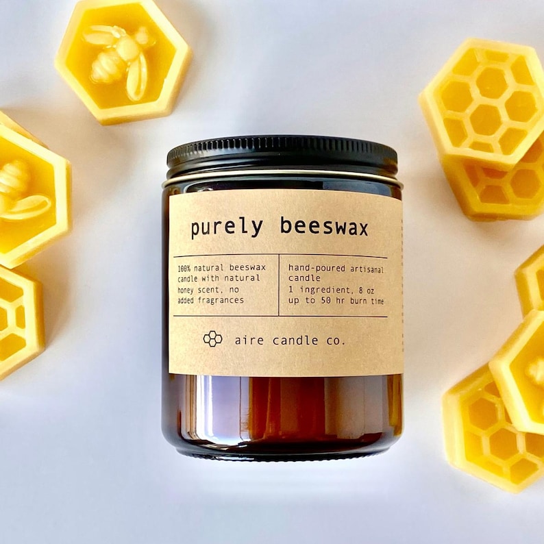 K&ouml;nnte beinhalten: Ein Glas-Kerze mit schwarzem Deckel, beschriftet mit "purely beeswax". Das Etikett beschreibt die nat&uuml;rlichen Inhaltsstoffe und den Honigduft der Kerze. Um die Kerze herum befinden sich gelbe Bienenwabenformen aus Bienenwachs. Die Kerze ist 227g.