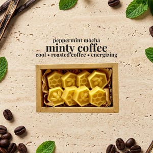 May include: A rectangular cardboard box containing eight yellow wax melts shaped like hexagons, featuring bee and honeycomb patterns. The text reads: peppermint mocha, minty coffee, cool, roasted coffee, energizing. Surrounded by coffee beans, mint leaves, and vanilla beans.