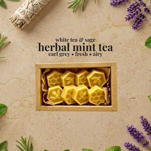 May include: A rectangular cardboard box filled with eight yellow hexagonal wax melts, each embossed with bee or honeycomb patterns. The text above reads white tea & sage herbal mint tea earl grey &bull; fresh &bull; airy, surrounded by sprigs of herbs.