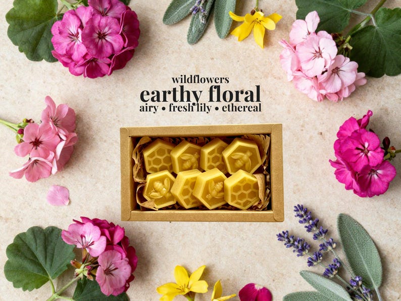 May include: A rectangular cardboard box containing eight yellow wax melts in hexagonal shapes, some embossed with bee and honeycomb patterns. The text above reads: wildflowers, earthy floral, airy, fresh lily, ethereal. Surrounded by pink flowers.