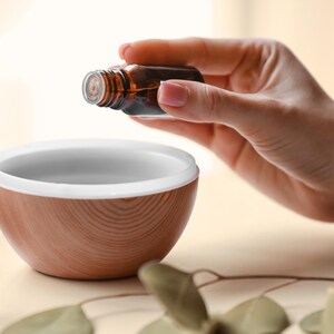 May include: A small amber glass bottle held above a round, wood-grain patterned bowl with a white interior rim. The bowl has a smooth, curved exterior surface featuring a light brown wood texture, while the glass bottle is dark brown and translucent.