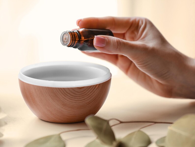May include: A small amber glass bottle held above a round, wood-grain patterned bowl with a white interior rim. The bowl has a smooth, curved exterior surface featuring a light brown wood texture, while the glass bottle is dark brown and translucent.