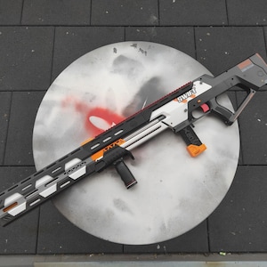 May include: A toy blaster gun in black, white, and orange, with the word "SWIFT" printed on the side. The gun features a long barrel with a patterned design and a handle. It rests on a circular, silver surface.