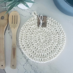 Puede incluir: Un posavasos de ganchillo blanco con una etiqueta marrón que dice "Tivet". El posavasos está sobre una superficie de mármol blanco con dos cucharas de madera.