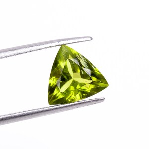 Op de afbeelding: Een driehoekige, gefacetteerde peridot edelsteen, vastgehouden door een zilveren pincet tegen een witte achtergrond. De peridot heeft een levendige, doorschijnende groene kleur, die de interne structuur en helderheid laat zien. De edelsteen is gesneden in een perfecte driehoek.