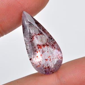 Puede incluir: Una piedra preciosa de cuarzo transparente en forma de lágrima con inclusiones rojas y plateadas. La piedra preciosa mide aproximadamente 5 cm de largo y se sostiene en una mano.