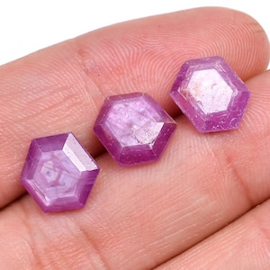 May include: Three faceted, hexagonal gemstones in shades of pink and purple. The gems are held in a hand, showcasing their polished surfaces and geometric cuts. The gems are likely rubies or sapphires.