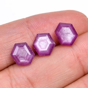 May include: Three faceted, hexagonal gemstones in a vibrant magenta hue. The gems are displayed against a neutral background, showcasing their geometric precision and polished surfaces. These gemstones are ideal for jewelry making or collecting.