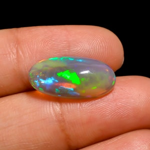 Può includere: Una gemma di opale ovale con un gioco di colori, che mostra vivaci lampi di verde, blu e arancione. La pietra è tenuta su uno sfondo scuro, evidenziando le sue qualità iridescenti. La superficie dell'opale è liscia e lucidata.