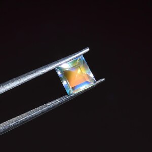May include: A square-cut gemstone held by metal tweezers, displaying a translucent appearance with iridescent flashes of orange, yellow, and blue light reflecting across its faceted surface against a solid black background.