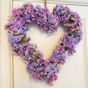 May include: A heart-shaped wreath constructed from dried statice flowers in shades of purple, lavender, and light blue. The floral arrangement is suspended by a thin, natural-coloured jute twine loop attached to the top centre of the heart structure.