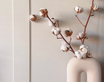 May include: A beige, U-shaped ceramic vase holds cotton stems with fluffy white blooms and brown seed pods. The vase sits on a wooden surface against a neutral-toned wall with vertical paneling. The arrangement creates a minimalist, natural aesthetic.