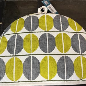 May include: A tea cosy featuring a repeating pattern of grey and chartreuse ovals on a white background. The ovals are divided by a vertical white line. A small white loop is at the top. Designed to keep tea warm.