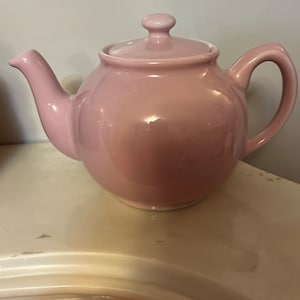 Può includere: Una teiera in ceramica rosa con un coperchio e un manico. La teiera è appoggiata su una superficie bianca.