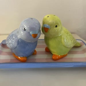 May include: A set of two ceramic bird salt and pepper shakers. The shakers are shaped like blue and yellow parakeets with orange beaks and feet. The shakers are sitting on a blue and pink striped tray.