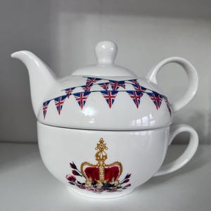 Può includere: Servizio da tè in ceramica bianca per una persona. Il coperchio della teiera presenta una ghirlanda di bandiere Union Jack. La tazza ha un design a corona bordato d'oro con dettagli floreali rossi e rosa.