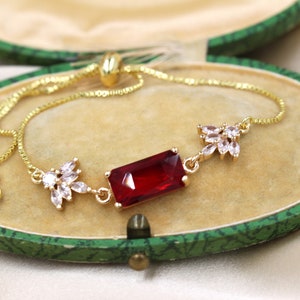 May include: A gold chain bracelet featuring a large red rectangular gemstone and two smaller clusters of clear rhinestones.