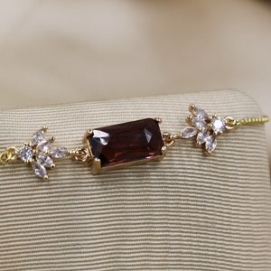Pode incluir: Uma pulseira de ouro com uma gema de cor púrpura escura e detalhes de cristal transparente. A pulseira tem uma corrente delicada e detalhes em forma de flor.