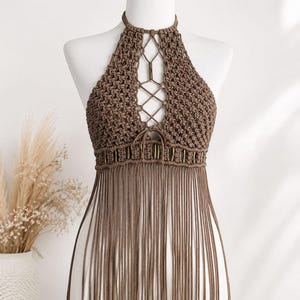 May include: A brown macrame halter top featuring a knotted lattice design, a lace-up front detail, and a row of metallic rings at the waist. The garment is finished with long, straight fringe hanging from the bottom hem, creating a textured look.