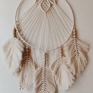 Puede incluir: Un atrapasueños de macramé blanco con cuentas de madera y detalles de plumas blancas.