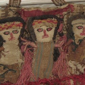 May include: Three small dolls made of woven fabric with fringe hair. The dolls are brown, tan, and beige with red and yellow accents. The dolls are arranged in a row and are sewn onto a larger piece of fabric.