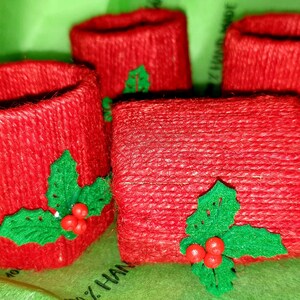 May include: Four red, handmade, cylindrical napkin rings with green holly leaf and red berry embellishments.