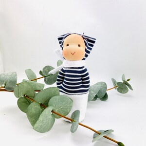 Könnte beinhalten: Eine handgefertigte Puppe mit einem gestreiften blau-weißen Outfit und einem weißen Hut. Die Puppe hat ein lächelndes Gesicht mit braunen Augen und einem braunen gestickten Mund. Die Puppe sitzt auf einer weißen Oberfläche mit grünen Blättern.