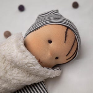 Könnte beinhalten: Eine handgefertigte Stoffpuppe mit einer gestreiften Mütze, braunen gestickten Haaren und einer weißen flauschigen Decke. Die Puppe liegt auf einer weißen Oberfläche.