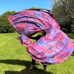 May include: Two large, flowing fabric flags in shades of pink, purple, and blue are held outdoors on a sunny day. The flags have a tie-dye pattern and are being waved in the air against a backdrop of green grass and trees.