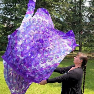 May include: A large, flowing, tie-dye fabric in shades of purple, white, and blue is held up in the air. The fabric has a square pattern in the center. The background is a green lawn and trees.