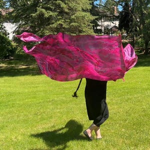 May include: A magenta silk scarf with a marbled pattern is held aloft. The scarf, in shades of pink and red, is rectangular and flowing. The person holding the scarf is wearing black trousers and shoes, standing on a green lawn.